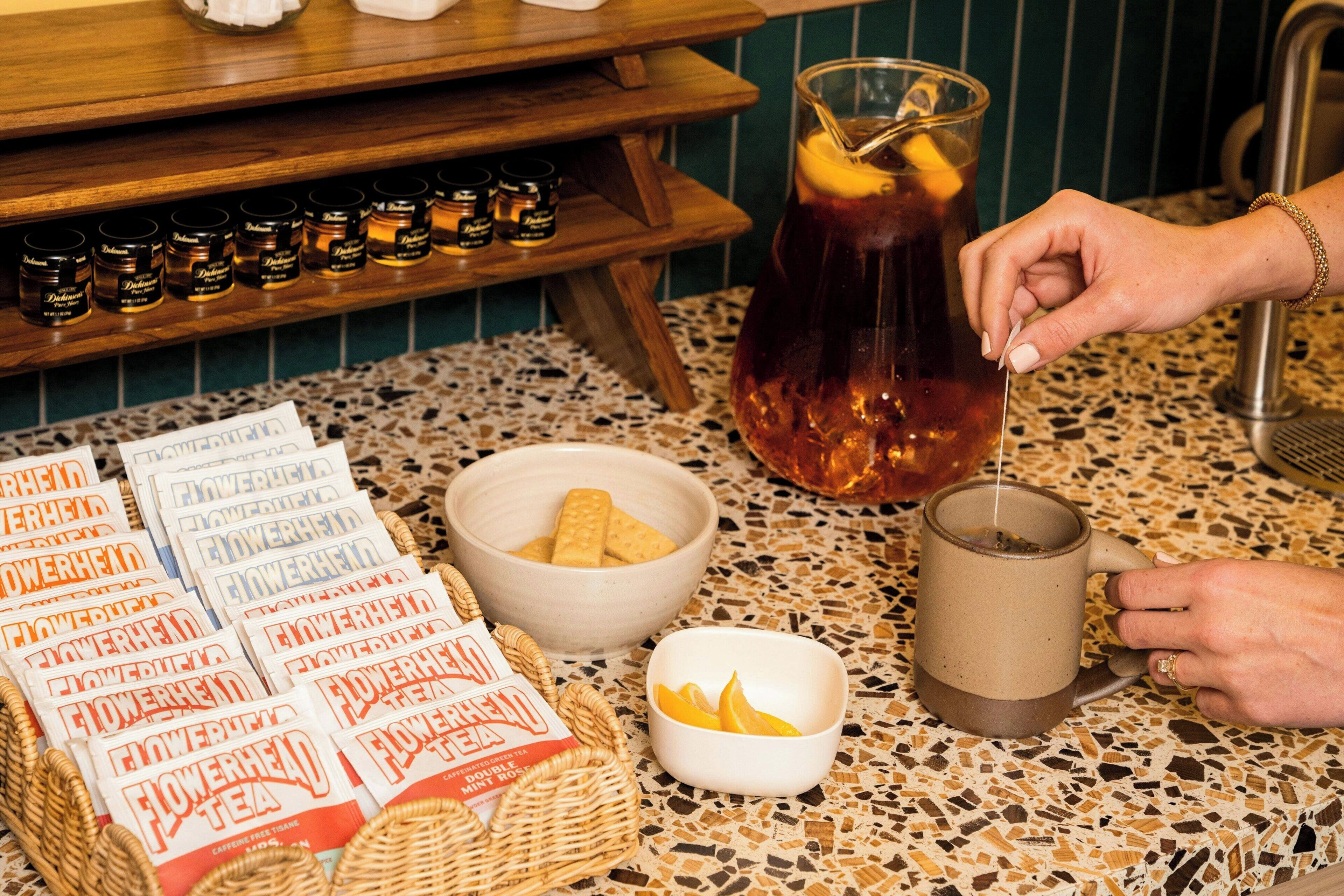 A countertop with a beverage pitcher, a mug, a bowl of crackers, a basket of tea packets, and a jar of honey.