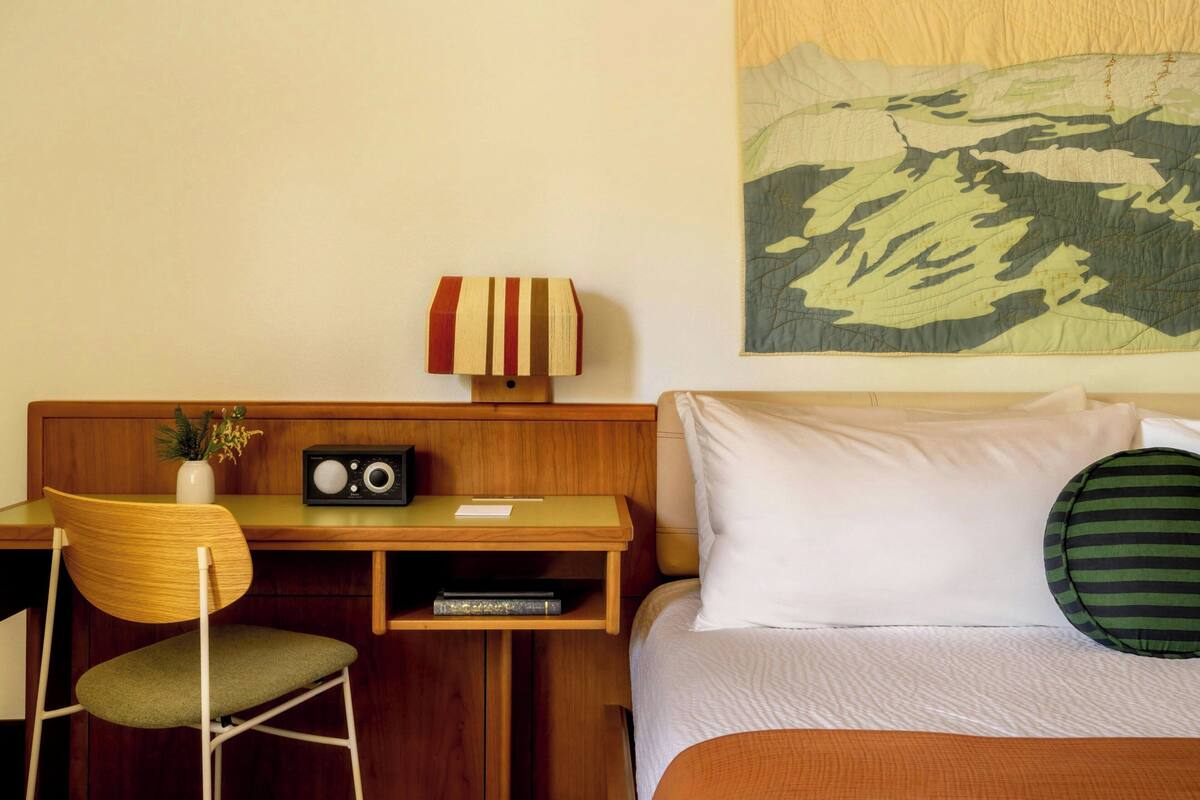 Une chambre d’hôtel avec un bureau en bois, une chaise, une radio, un vase avec des fleurs, un lit avec une literie blanche et une grande tapisserie murale.