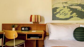 A hotel room with a wooden desk, a chair, a radio, a vase with flowers, a bed with white bedding, and a large wall tapestry.