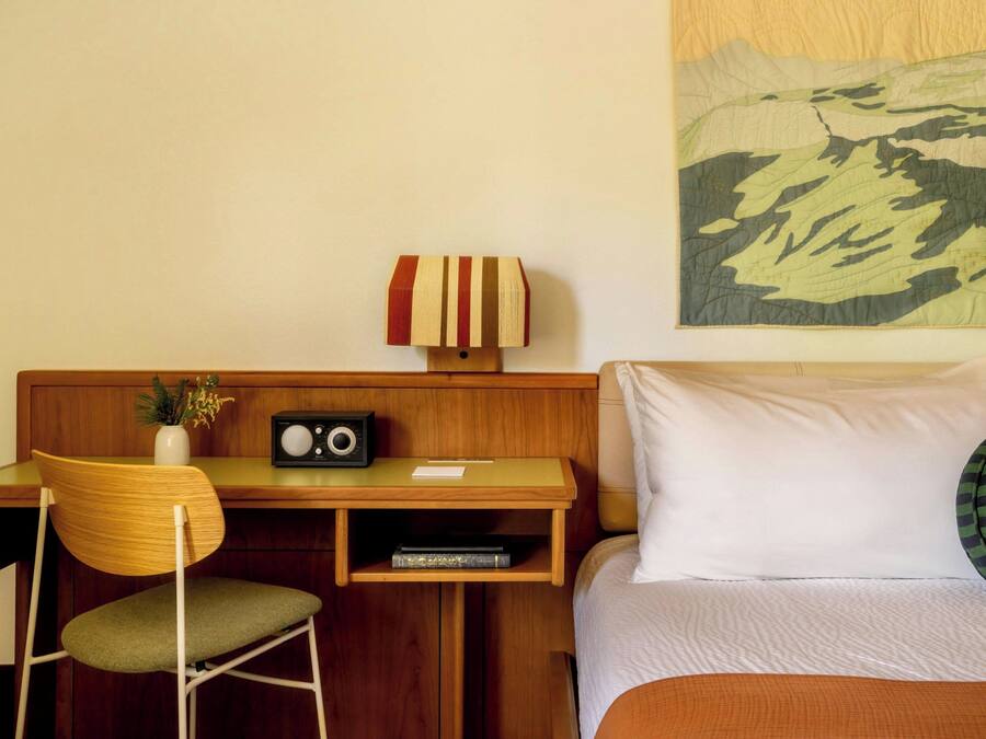 A hotel room with a wooden desk, a chair, a radio, a vase with flowers, a bed with white bedding, and a large wall tapestry.