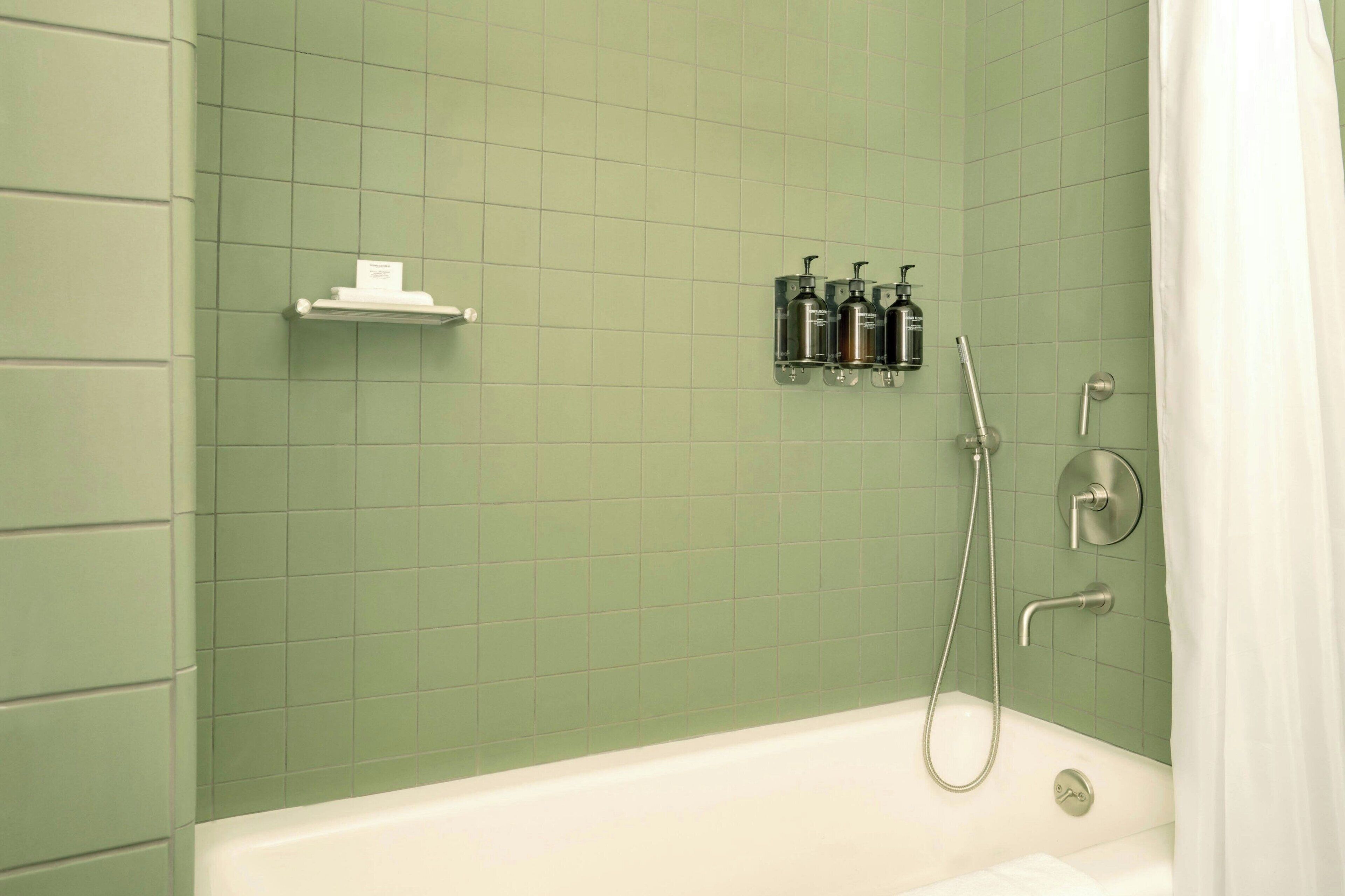 A bathroom with a green tiled wall, a white bathtub, a shower with a handheld showerhead, and a shelf with toiletries.