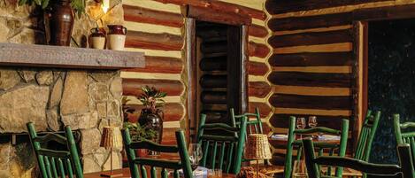 A rustic wooden cabin interior with a dining table set for four, surrounded by green chairs, a fireplace, and a stone wall.
