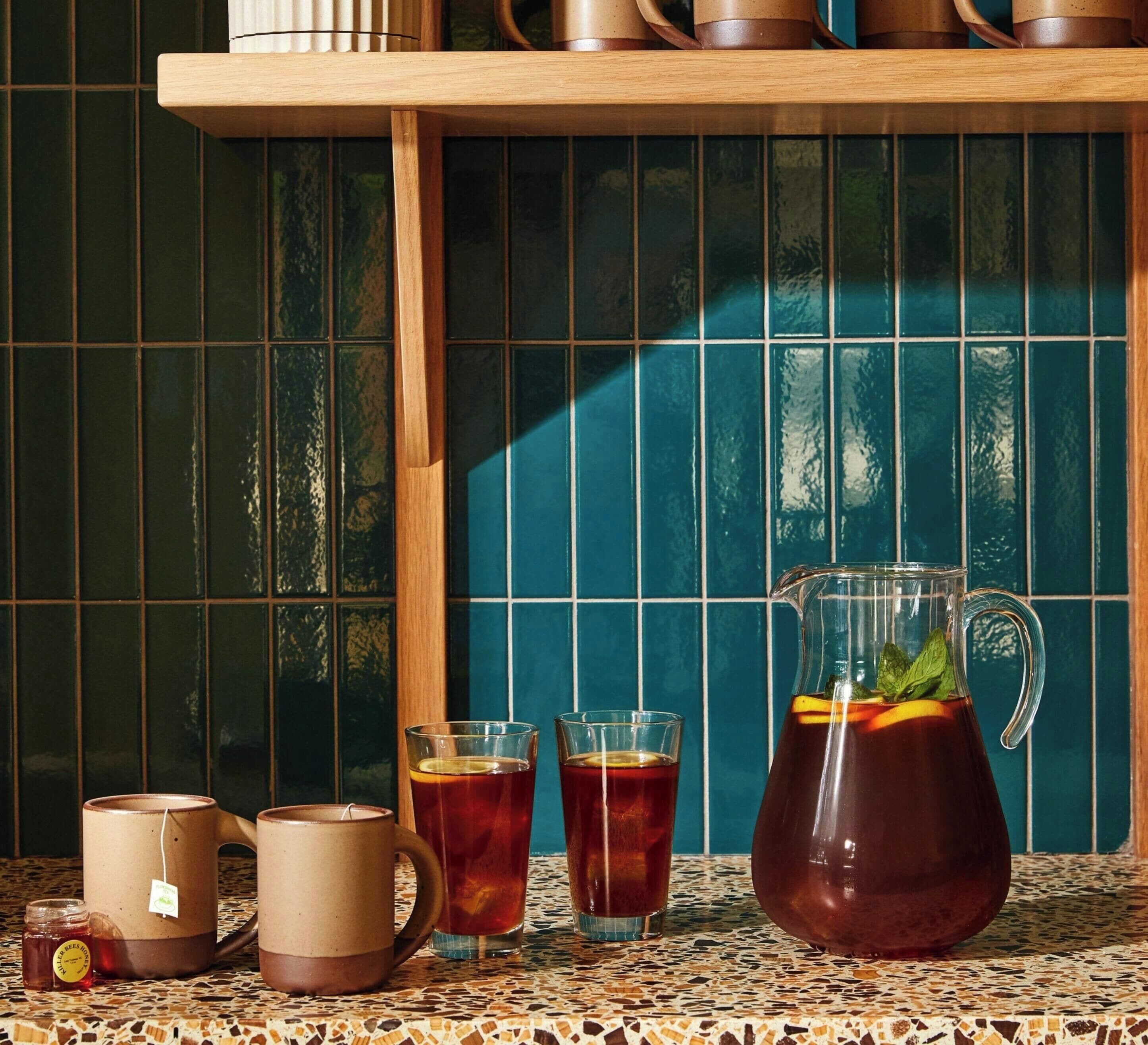 A kitchen counter with a pitcher of iced tea, two mugs, and a jar of honey.