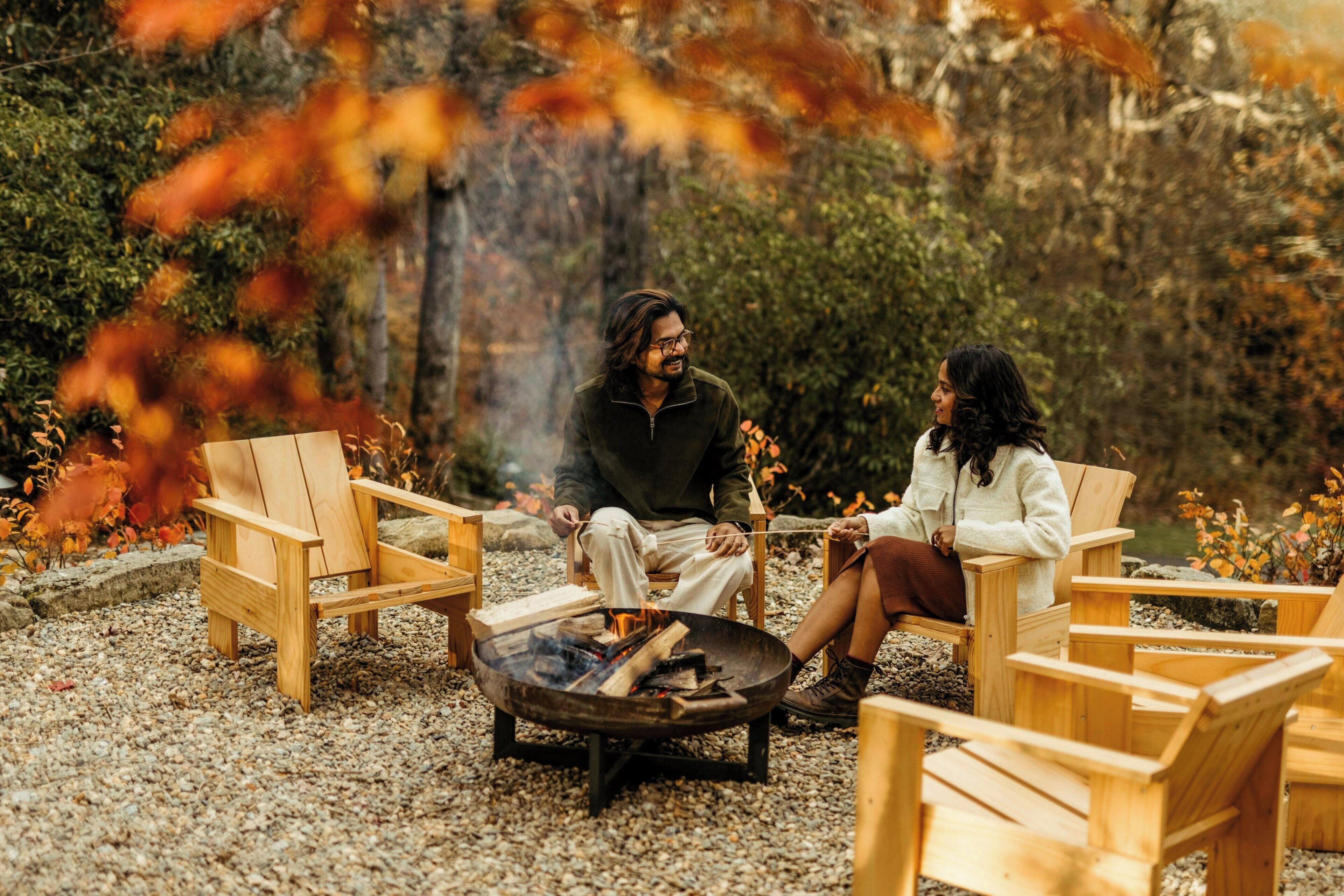 Two people sitting on wooden chairs around a campfire in a natural setting with trees and a rocky ground.