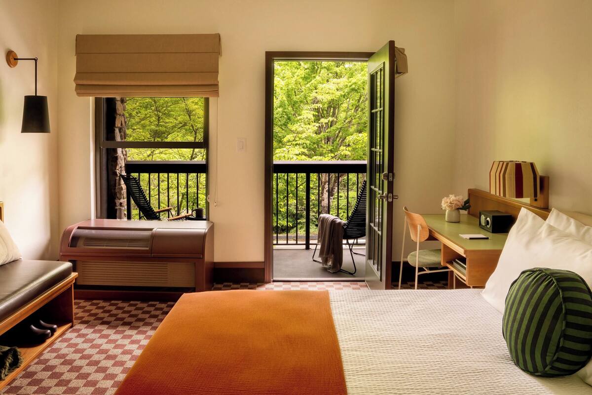 Une chambre d’hôtel comprenant un lit, un bureau avec une chaise, une petite table avec un vase, un radiateur, une fenêtre donnant sur un espace verdoyant et une porte menant à un balcon extérieur.