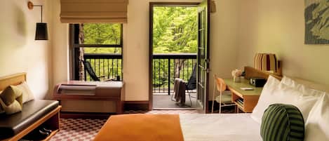A hotel room with a bed, a desk with a chair, a small table with a vase, a radiator, a window with a view of greenery, and a door leading to an outdoor balcony.