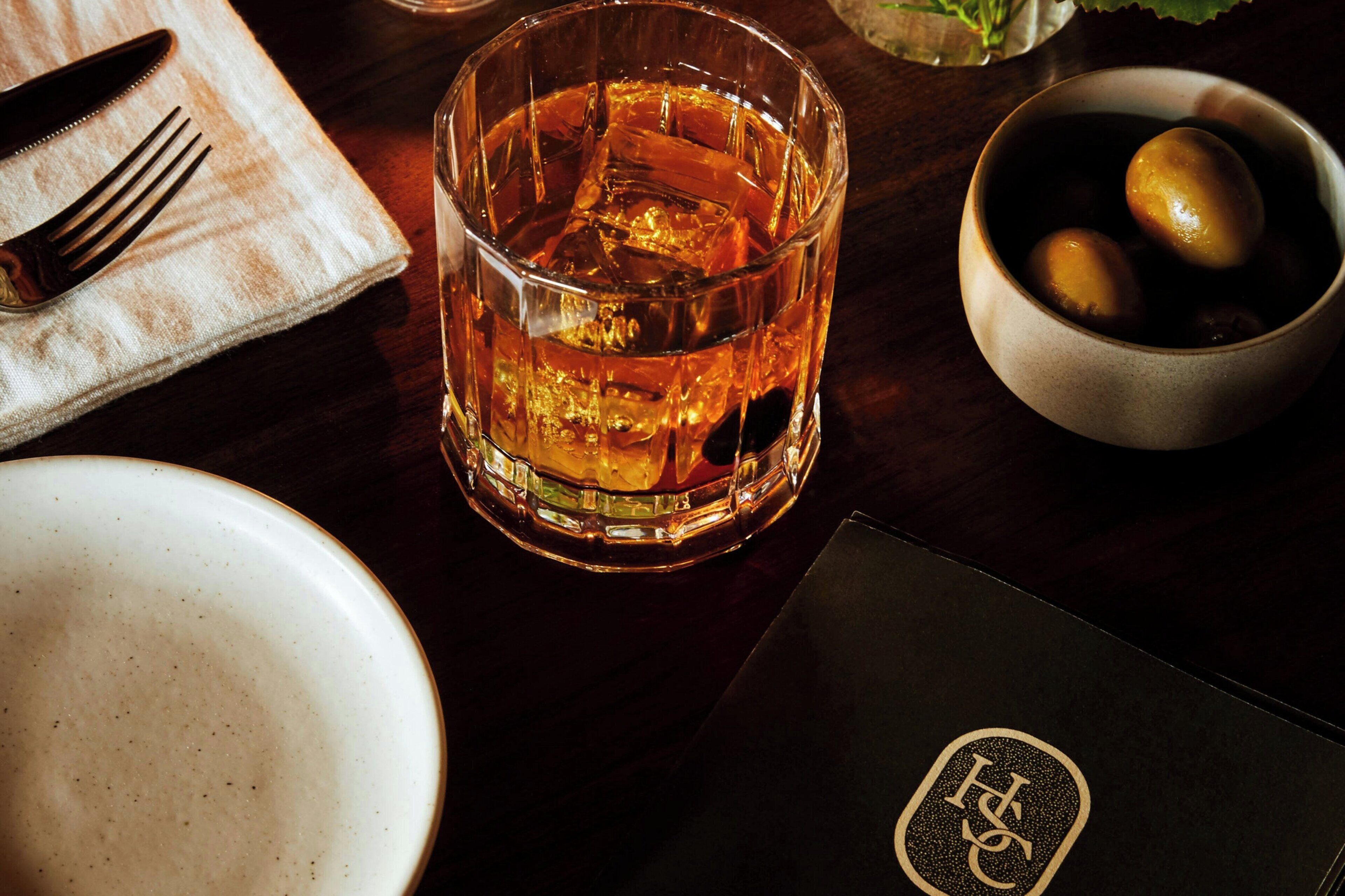 Een glas whiskey, een schaal met citroenen, een servet en een menukaart op een houten tafel.
