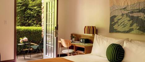 Une chambre d’hôtel avec un lit, un bureau, une chaise et une vue sur un jardin, accessible par une porte ouverte.