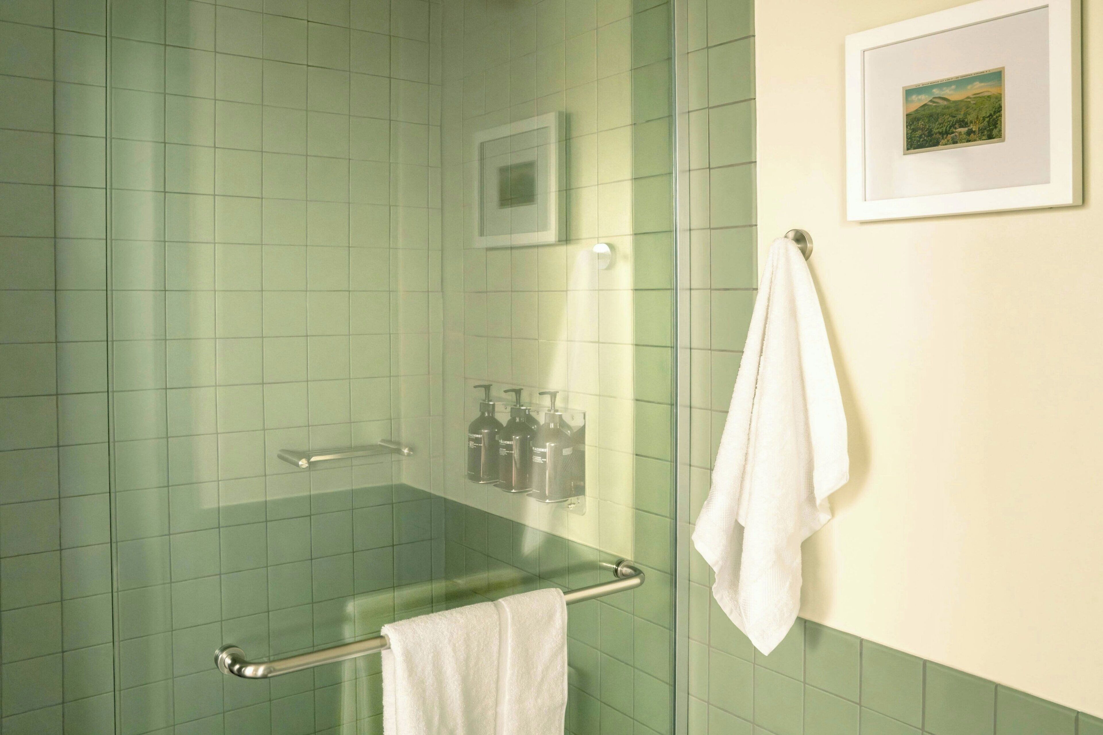 A bathroom with green tiles, a glass shower enclosure, a white towel on a rack, and a framed picture on the wall.