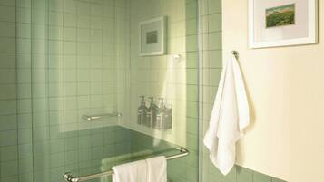 A bathroom with green tiles, a glass shower enclosure, a white towel on a rack, and a framed picture on the wall.
