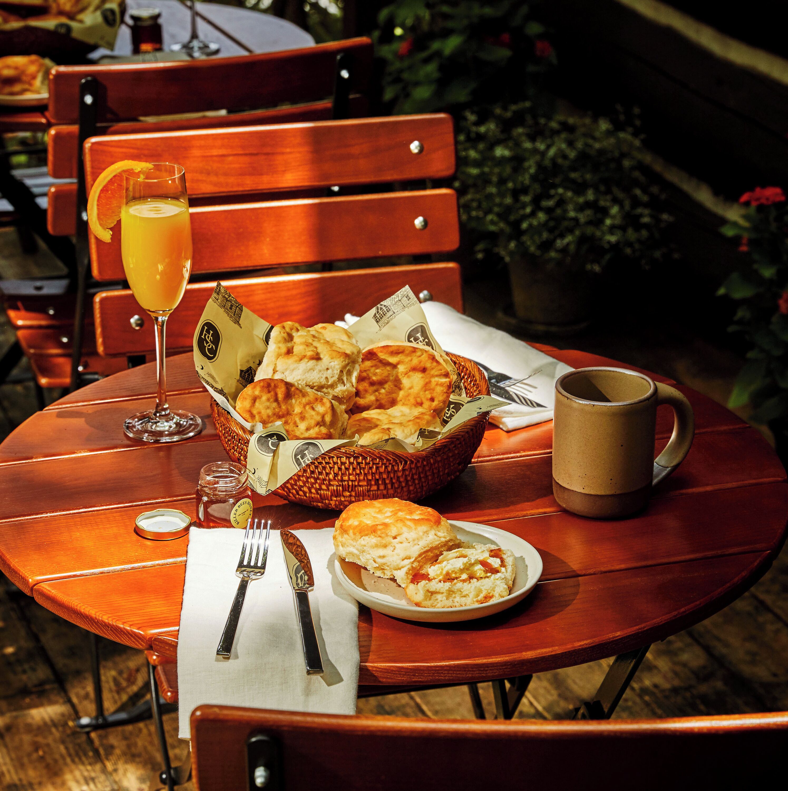 A wooden table with a basket of biscuits, a glass of orange juice, a plate with a biscuit sandwich, a mug, and cutlery on a napkin.