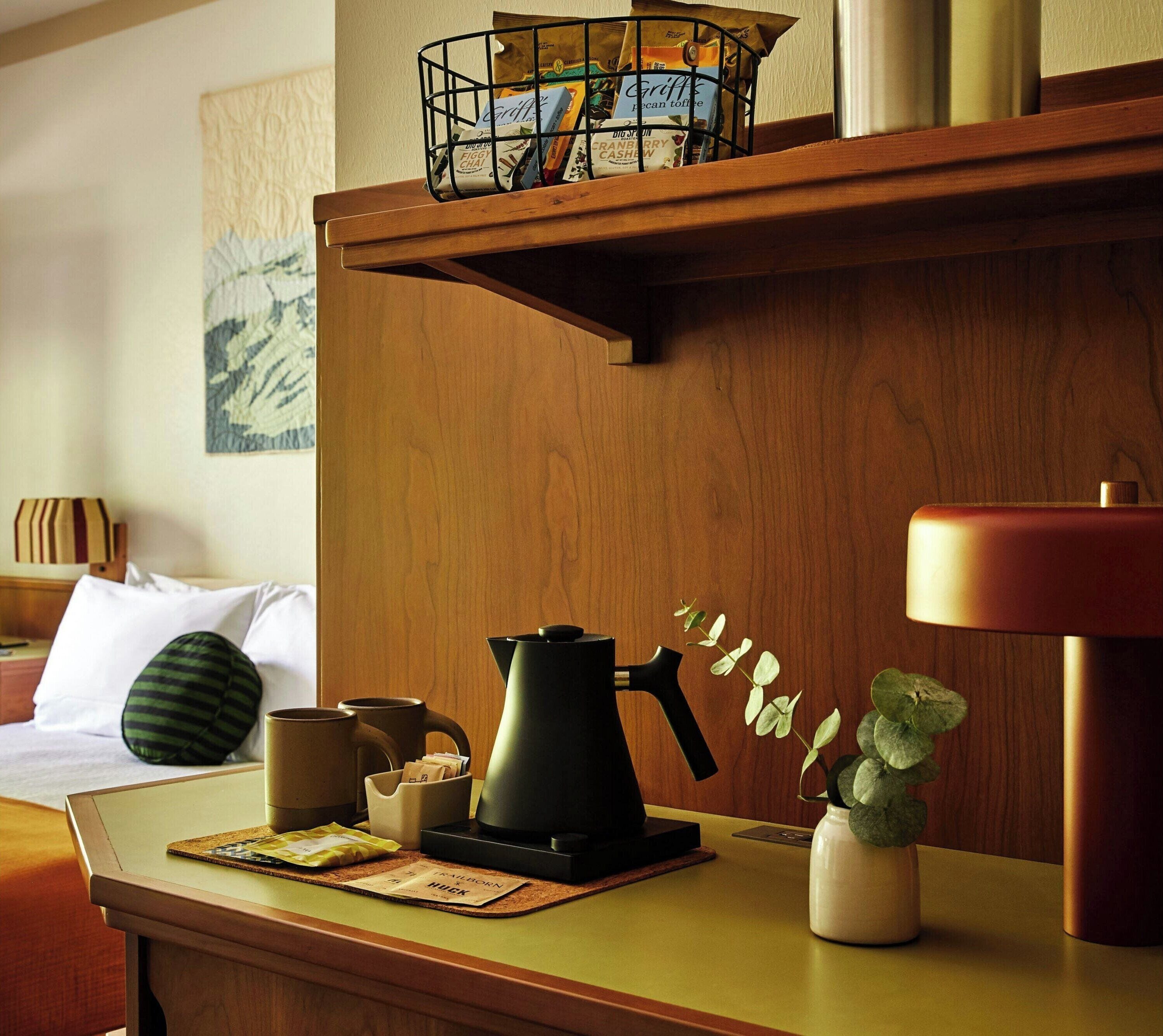 A hotel room with a wooden desk, a coffee maker, a kettle, a mug, a plant, and a wall-mounted shelf with a basket containing coffee bags.