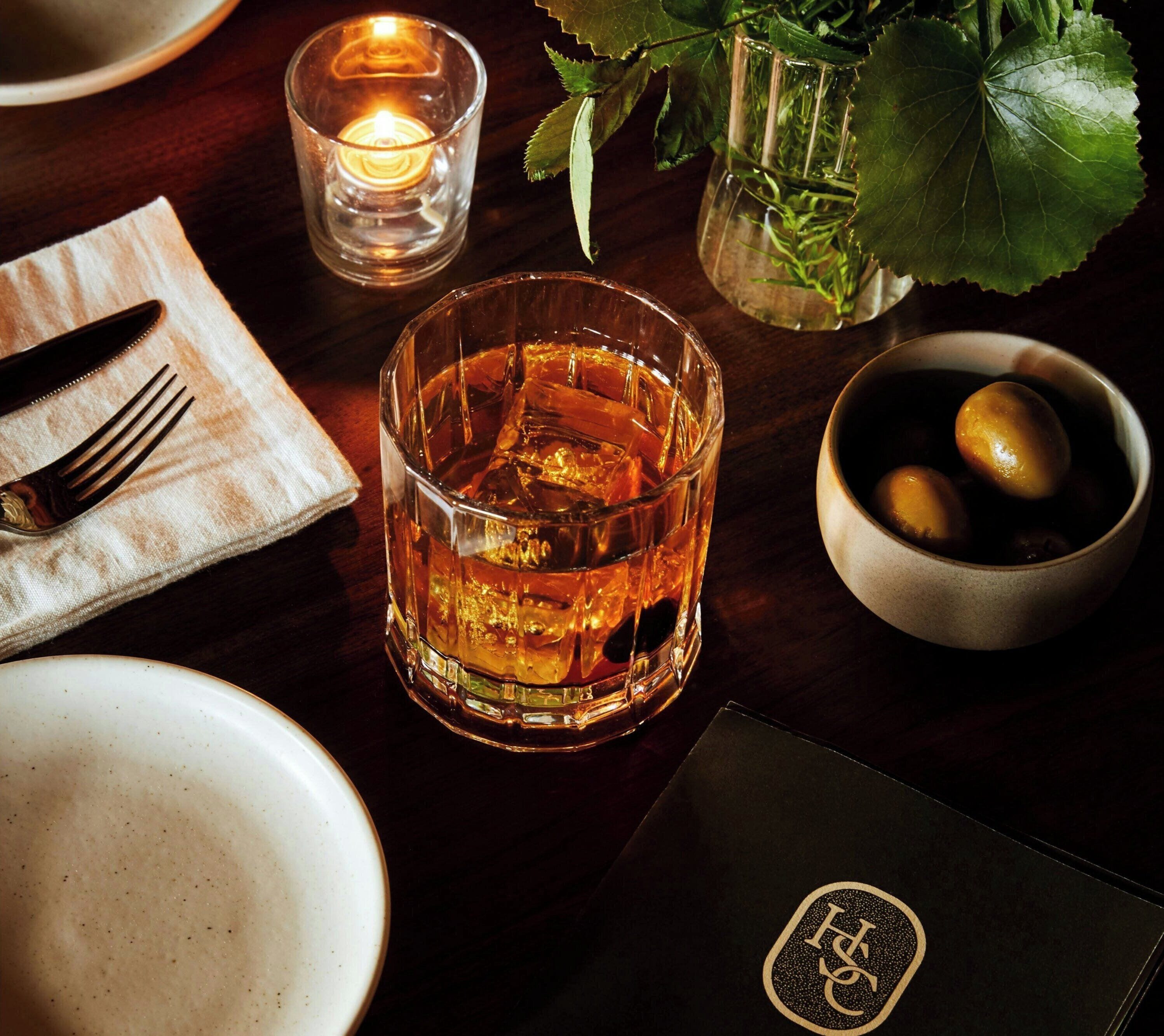 A table setting with a glass of whiskey, a candle, a bowl of olives, and a menu.