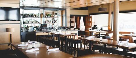 A restaurant with multiple tables and chairs, a bar area with shelves of bottles, and large windows allowing natural light.
