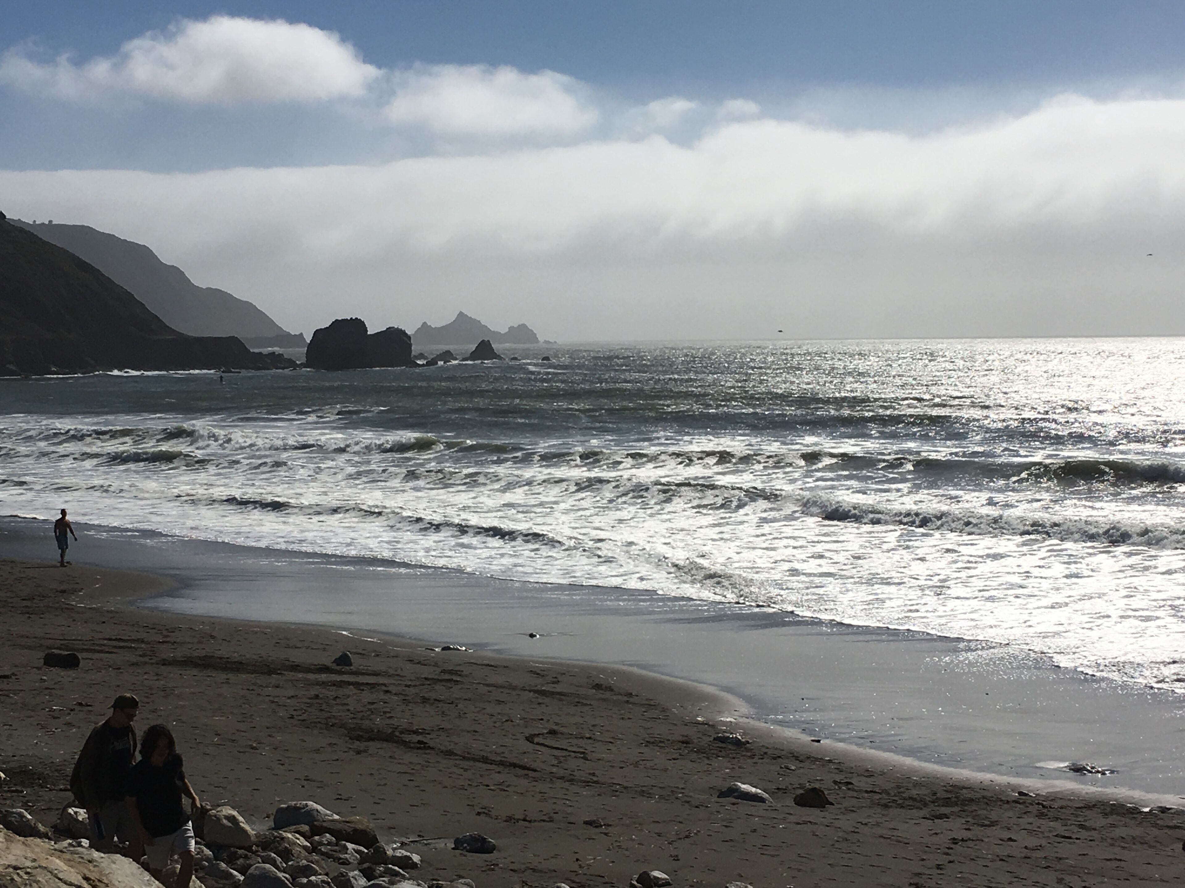 Una playa con olas, rocas y montañas al fondo.