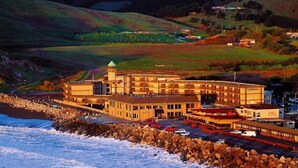A coastal hotel with a prominent tower, situated by the sea with rocky shorelines and hilly terrain in the background. - Pacifica Lighthouse Hotel Trademark Collection by Wyndham (Pacifica)