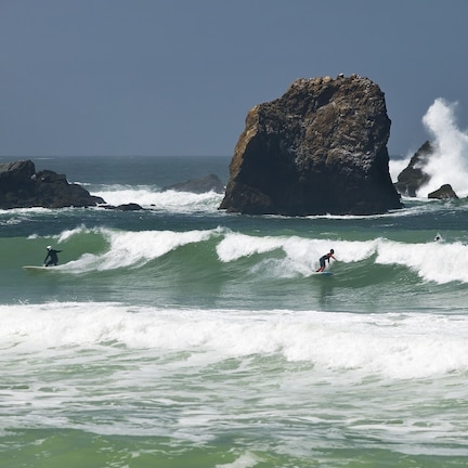 A coastal scene with large rock formations and surfers riding waves.