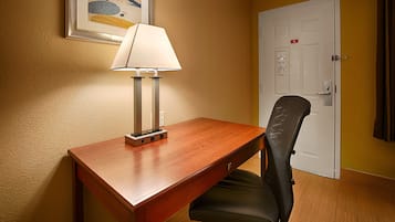 Habitación de hotel con un escritorio de madera, una silla de malla negra, una puerta blanca y un cuadro en la pared.