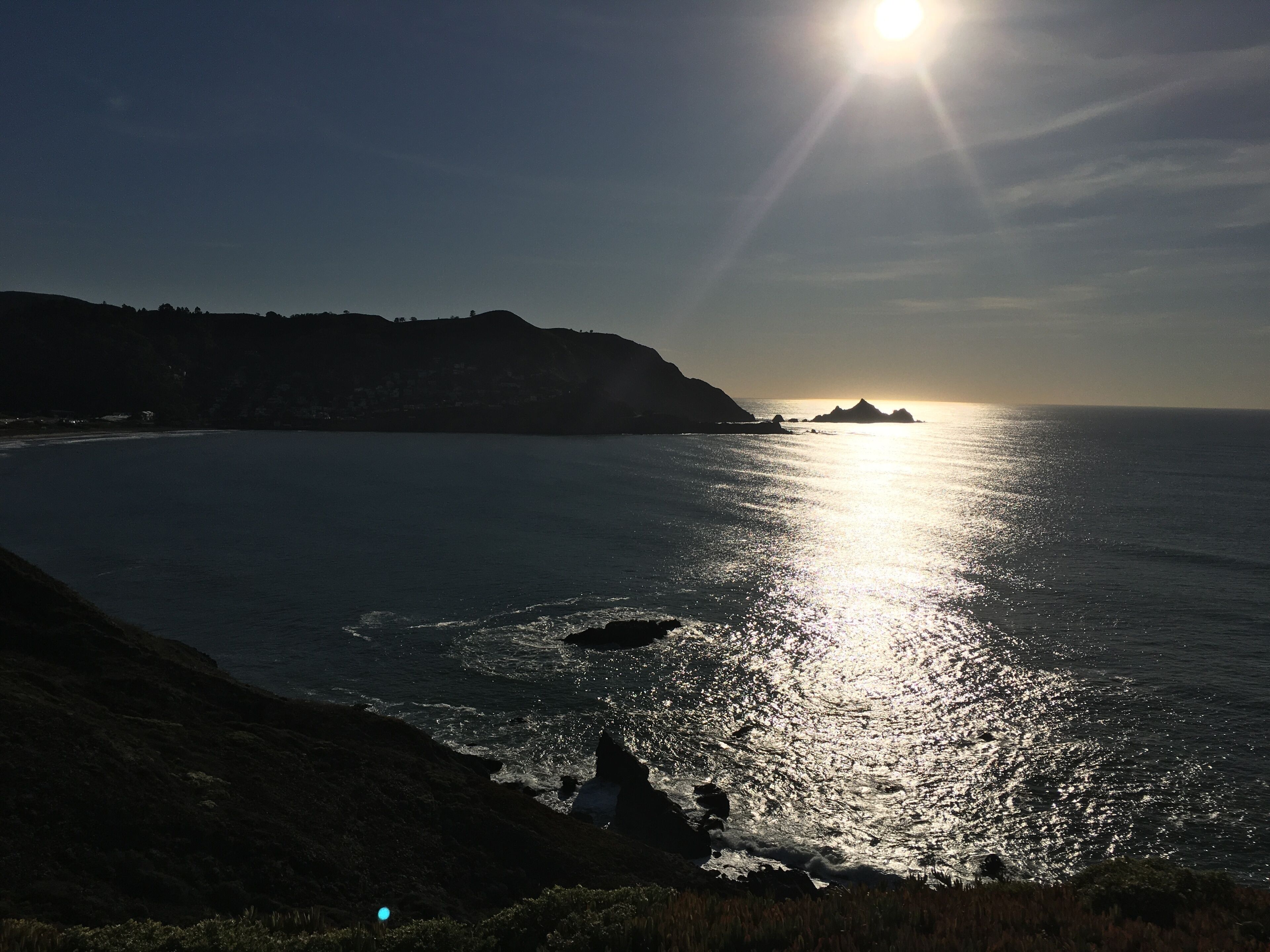 Un paisaje costero con cielo despejado, el sol reflejándose en el agua y una masa de tierra a lo lejos.