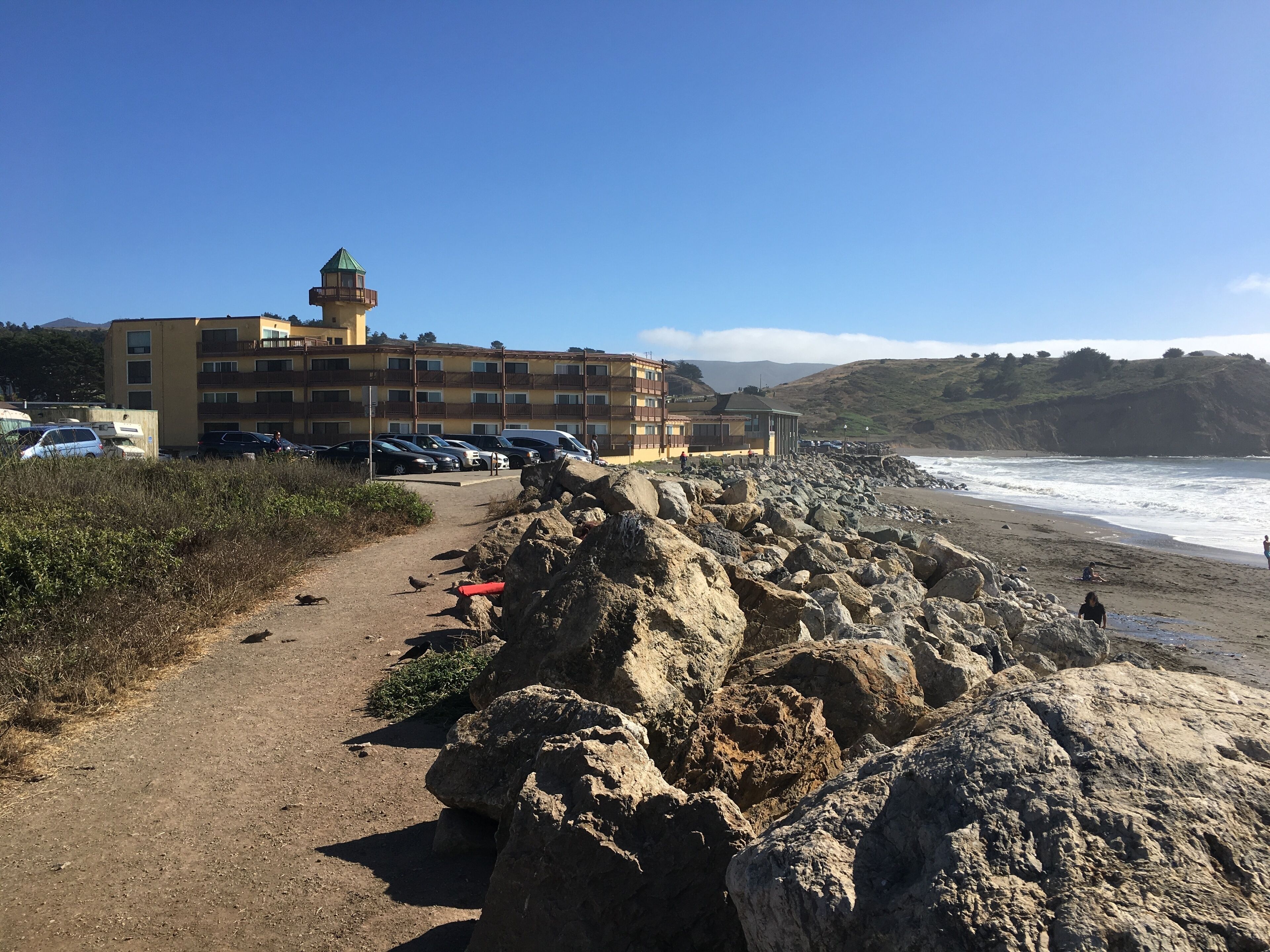 A coastal hotel with a rocky walkway leading to the beach, a parking area, and a view of the ocean.