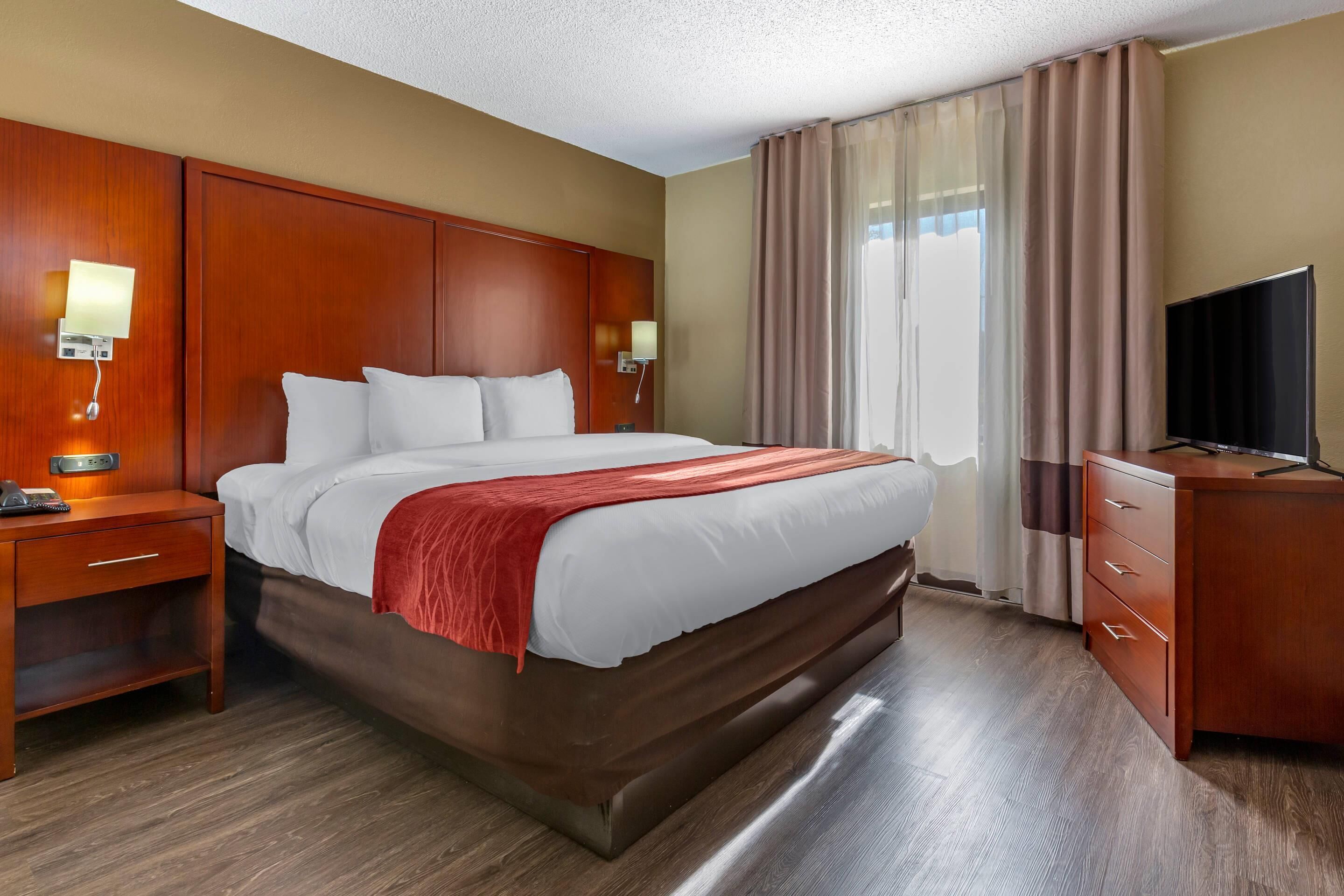 A hotel room with a large bed, wooden headboard, bedside lamps, a desk with a television, and a window with curtains.