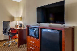 A hotel room with a flat-screen TV, a microwave, a refrigerator, a desk with a chair, and a lamp.