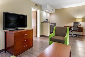 A hotel room with a wooden dresser, a television, a green armchair, a desk with a chair, and a small kitchenette.