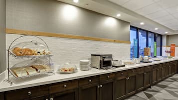 Een ontbijtruimte van een hotel met een buffet, inclusief diverse gebakjes en belegde broodjes, een koffieapparaat en een drankstation voor zelfbediening.