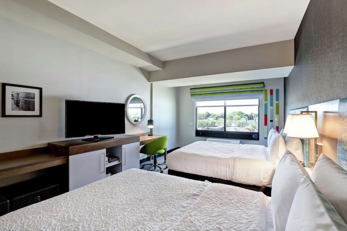 Habitación de hotel con dos camas, un escritorio con televisor de pantalla plana, una silla verde y una ventana con vista a áreas verdes.