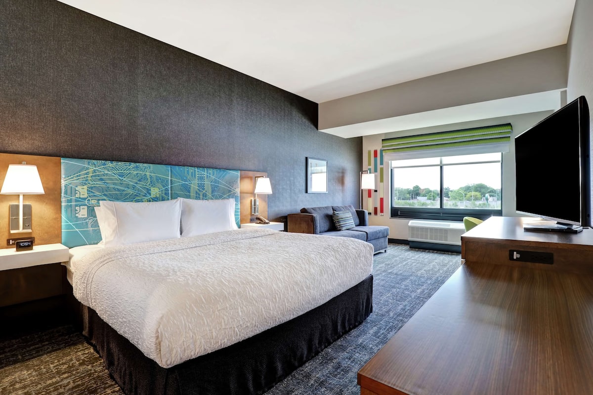 Habitación de hotel moderna con una cama grande, un sofá, un televisor y una ventana con vistas a la ciudad.