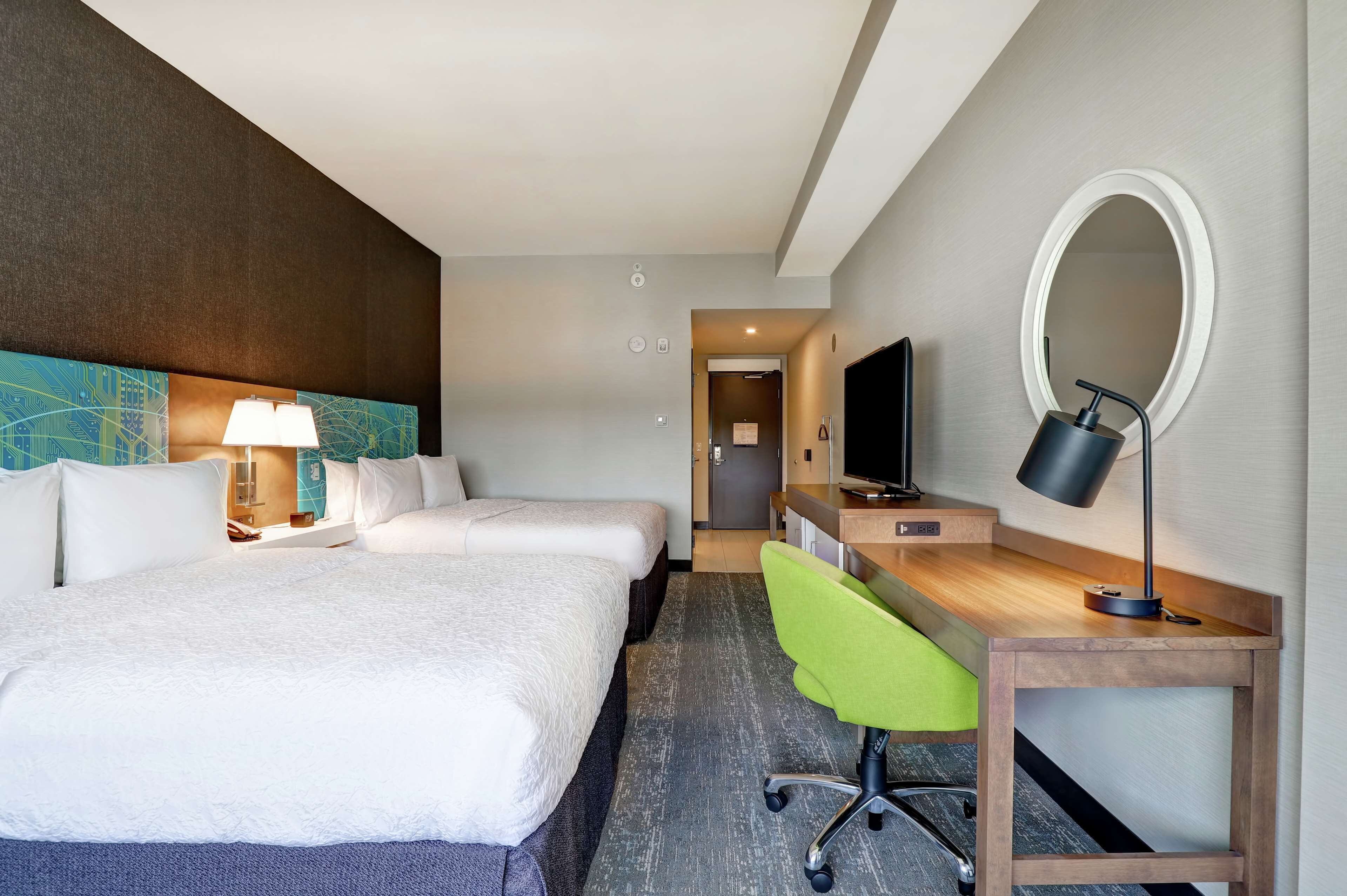 Una habitación de hotel con dos camas, un escritorio con silla, un televisor, un espejo y una lámpara.