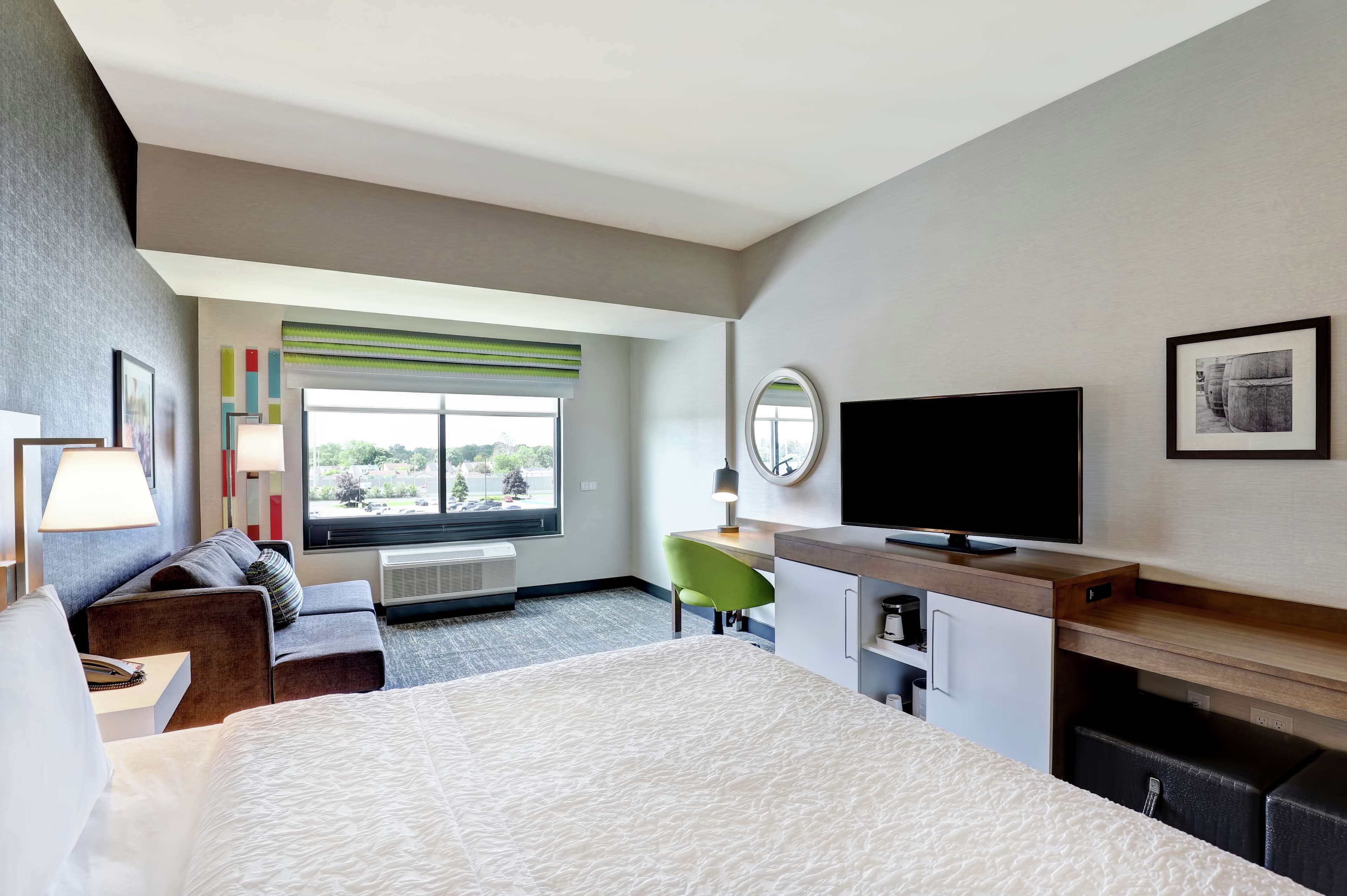 Habitación de hotel moderna con una cama grande, un escritorio, un televisor y una ventana con vistas a zonas verdes.