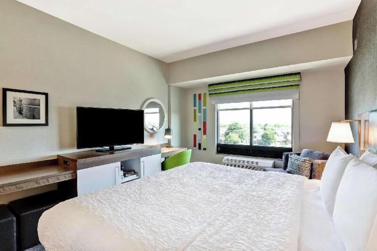 A hotel room with a large bed, a flat-screen TV, a desk with a chair, a window with a view, and a wall-mounted mirror.