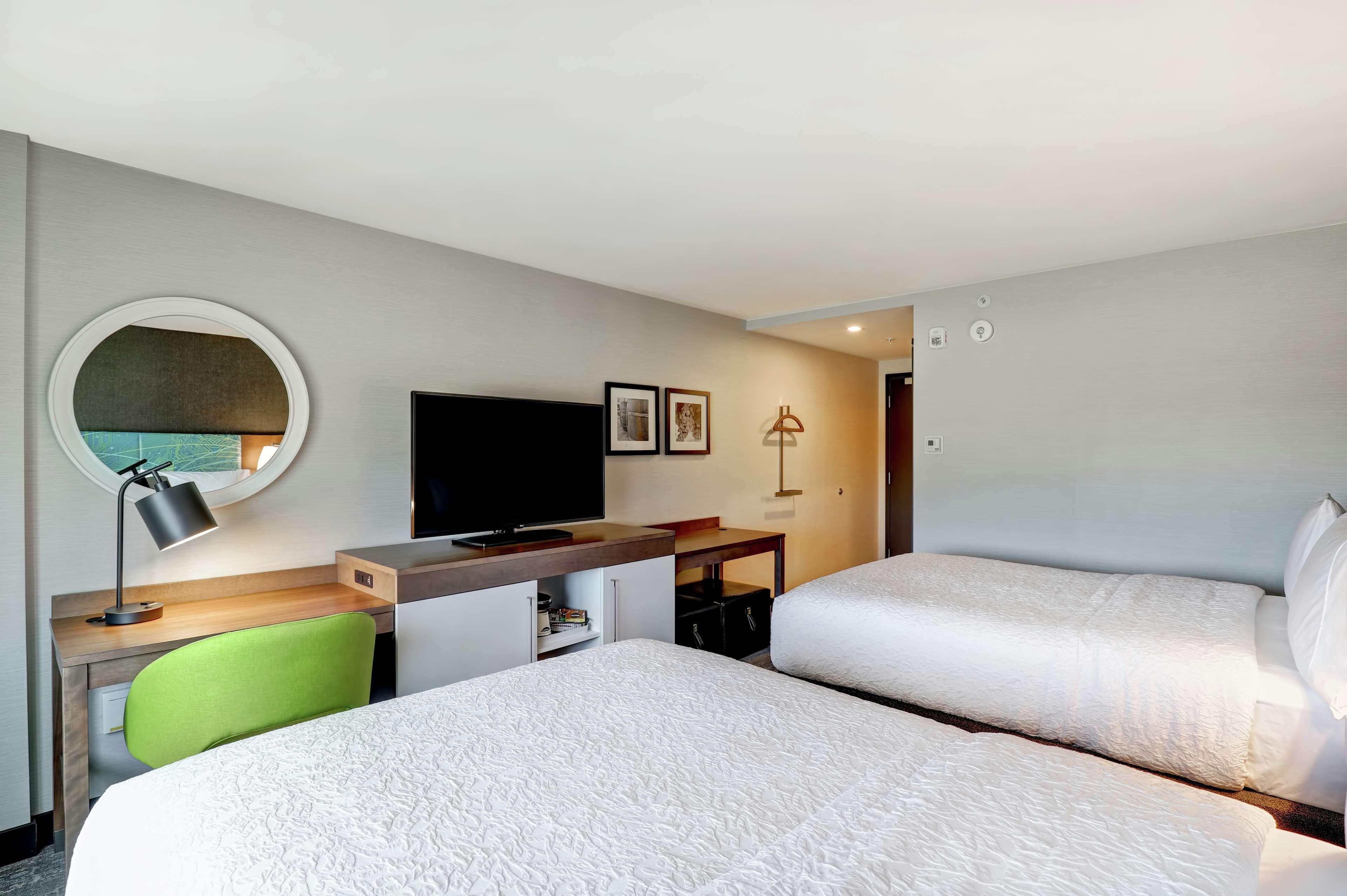 Quarto de hotel com duas camas, uma escrivaninha com computador, televisão e espelho redondo.
