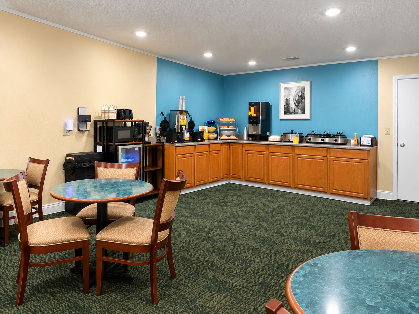 A hotel room with a kitchenette, a dining area with a round table and chairs, and a blue accent wall.
