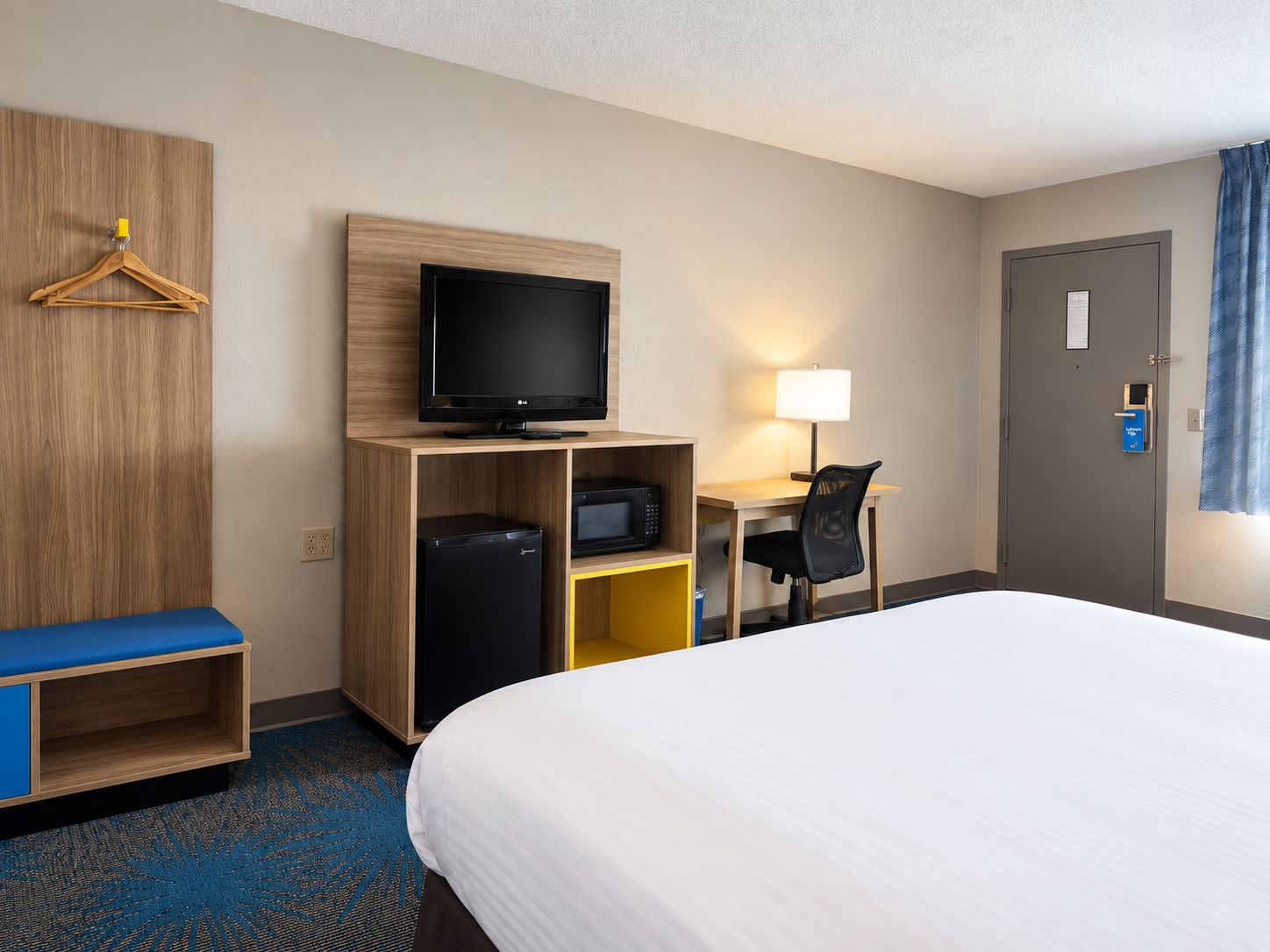 A hotel room with a bed, a desk with a lamp, a television, a microwave, a mini-fridge, and a hanging coat rack.