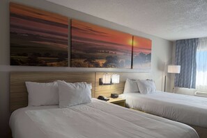 Een hotelkamer met twee bedden, een houten hoofdeinde, een nachtkastje met telefoon en lamp, en een groot schilderij van een zonsondergang boven de bedden.