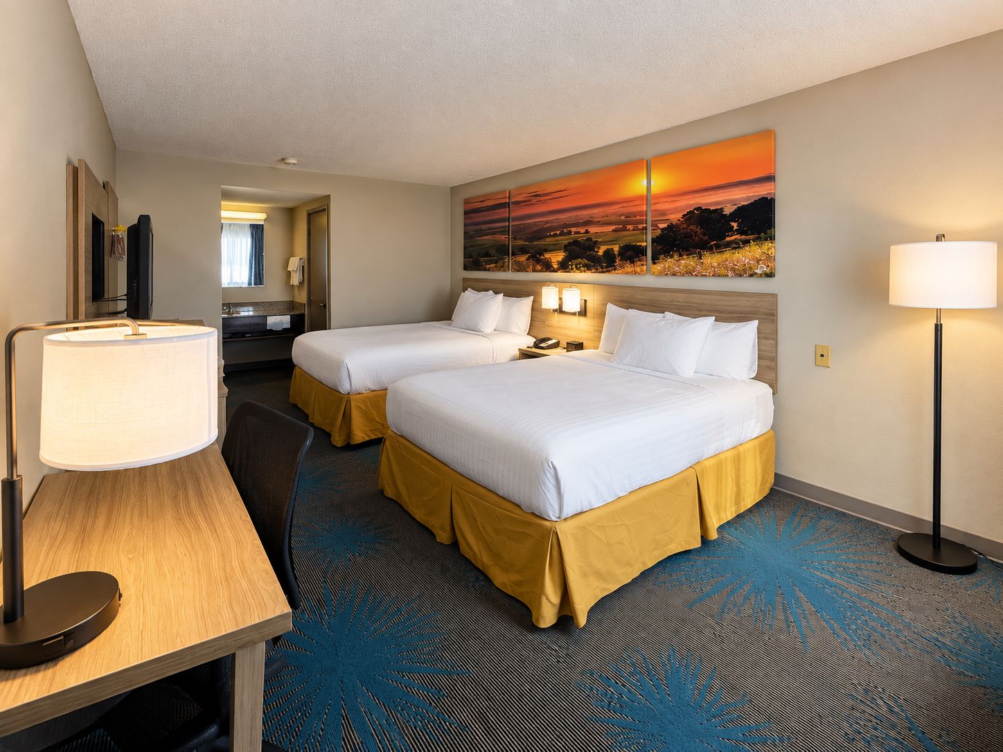 A hotel room with two beds, a desk, a lamp, a painting, and a carpet.