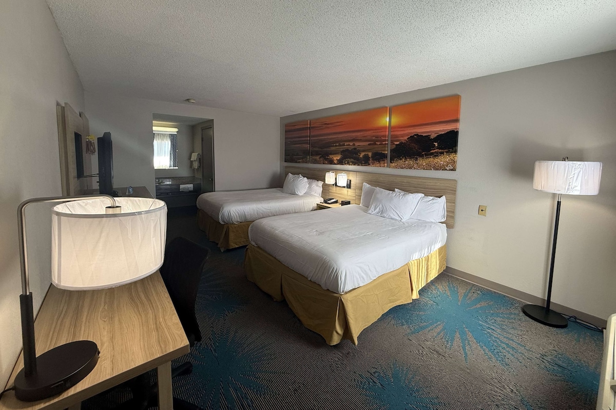 Habitación de hotel con dos camas, un escritorio, una lámpara y un cuadro de un atardecer.