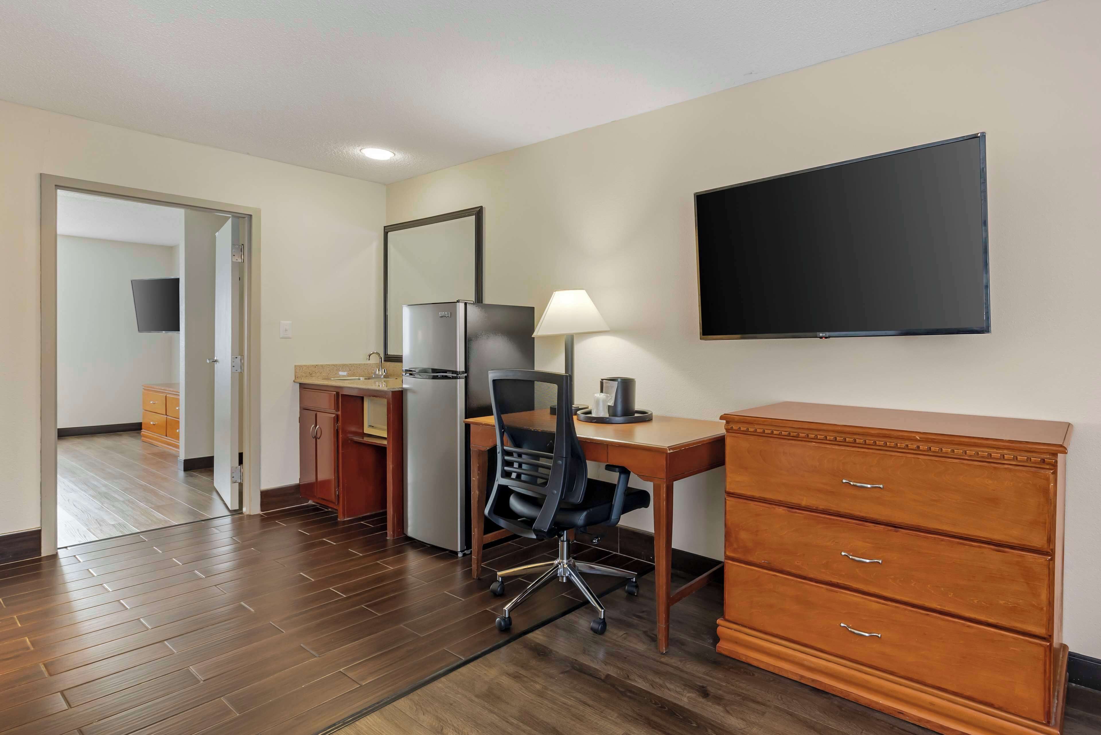 Une chambre d’hôtel équipée d’un bureau en bois, d’une chaise, d’une commode, d’un téléviseur à écran plat, d’un réfrigérateur et d’un coin cuisine avec des placards et un four à micro-ondes.