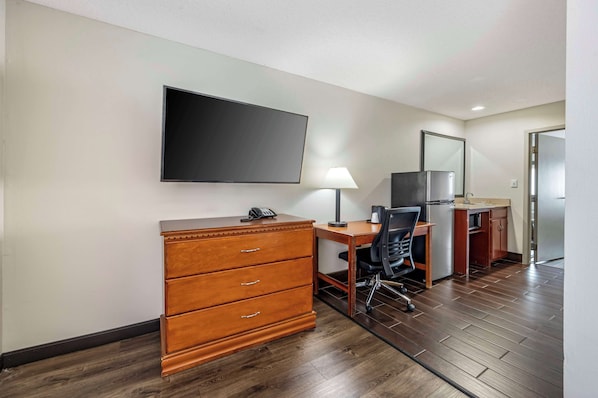 A hotel room with a wooden desk, a chair, a flat-screen TV, a dresser, a lamp, and a refrigerator.