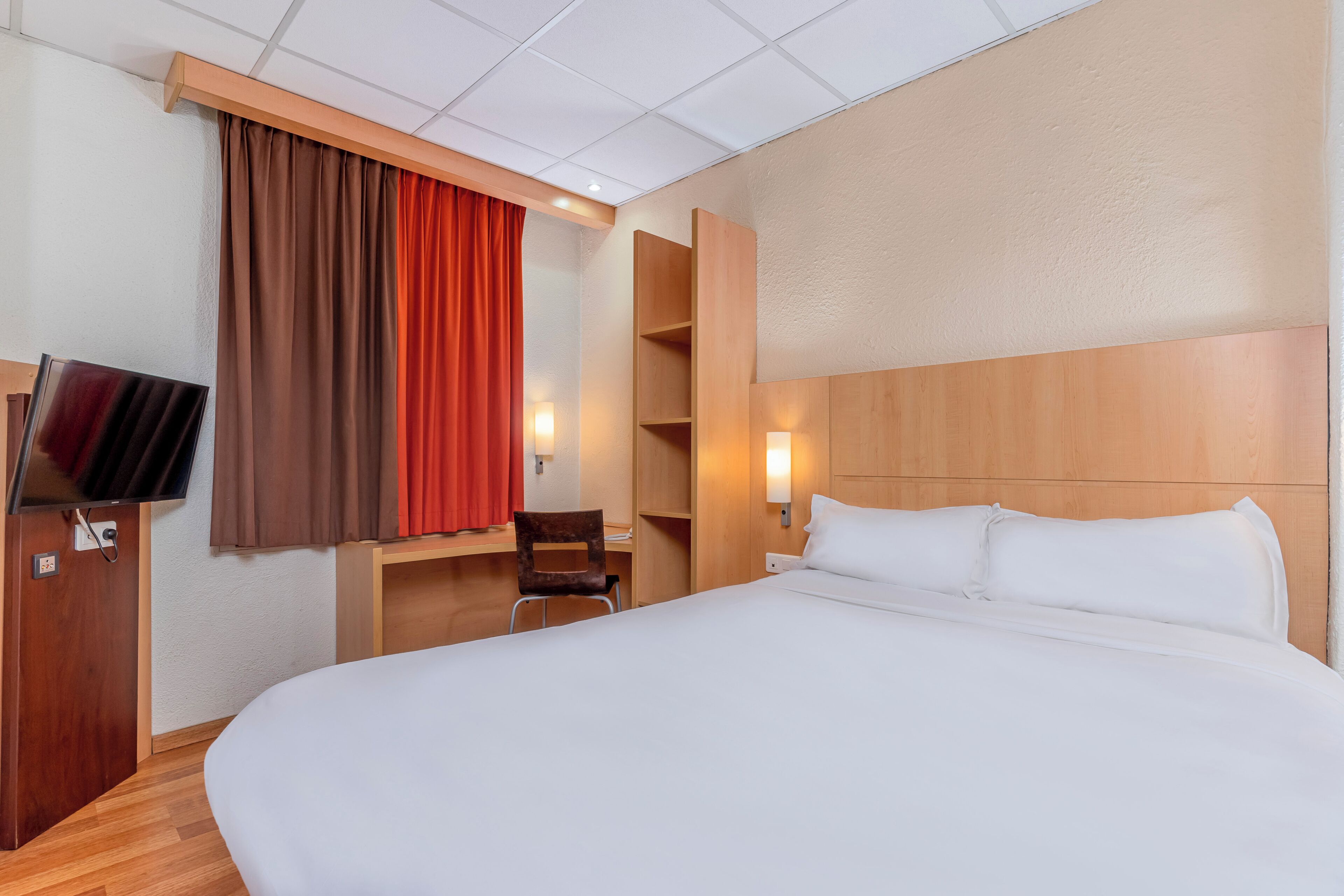 A hotel room with a large bed, a desk, a chair, a TV, and a wardrobe.