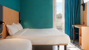 Habitación de hotel con cama, escritorio, silla, ventana con cortinas y vistas a edificios.