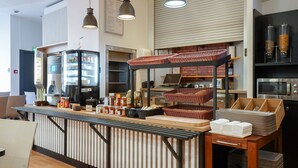 Un área de cocina moderna con una barra que exhibe alimentos, una cafetera y un microondas. Hay cestas y recipientes en la barra, y una mesa de madera con platos apilados.
