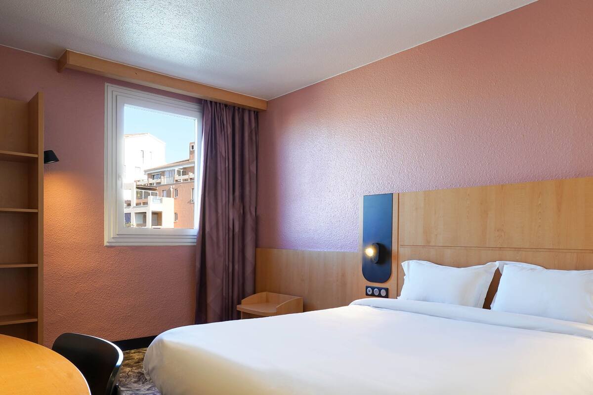 A hotel room with a bed, a desk, a chair, a window with curtains, and a view of buildings outside.