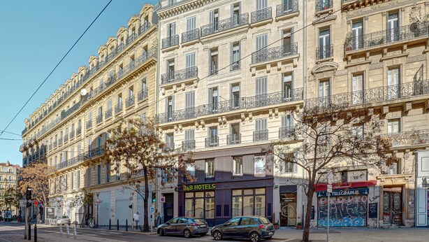 Un hotel de varios pisos con balcones, una fachada con rótulos y automóviles estacionados.