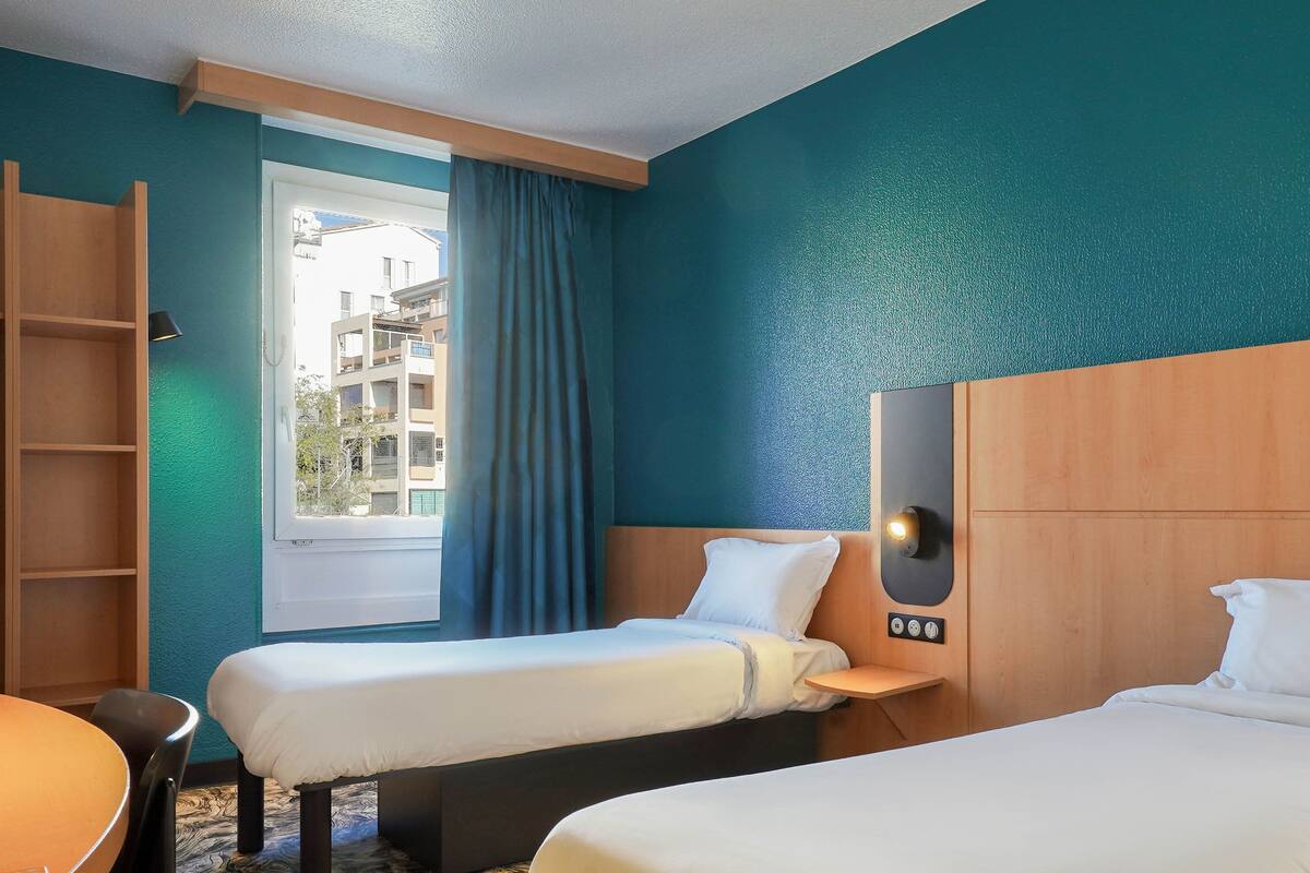 A hotel room with two single beds, a wooden headboard, a window with curtains, a small table, and a chair.
