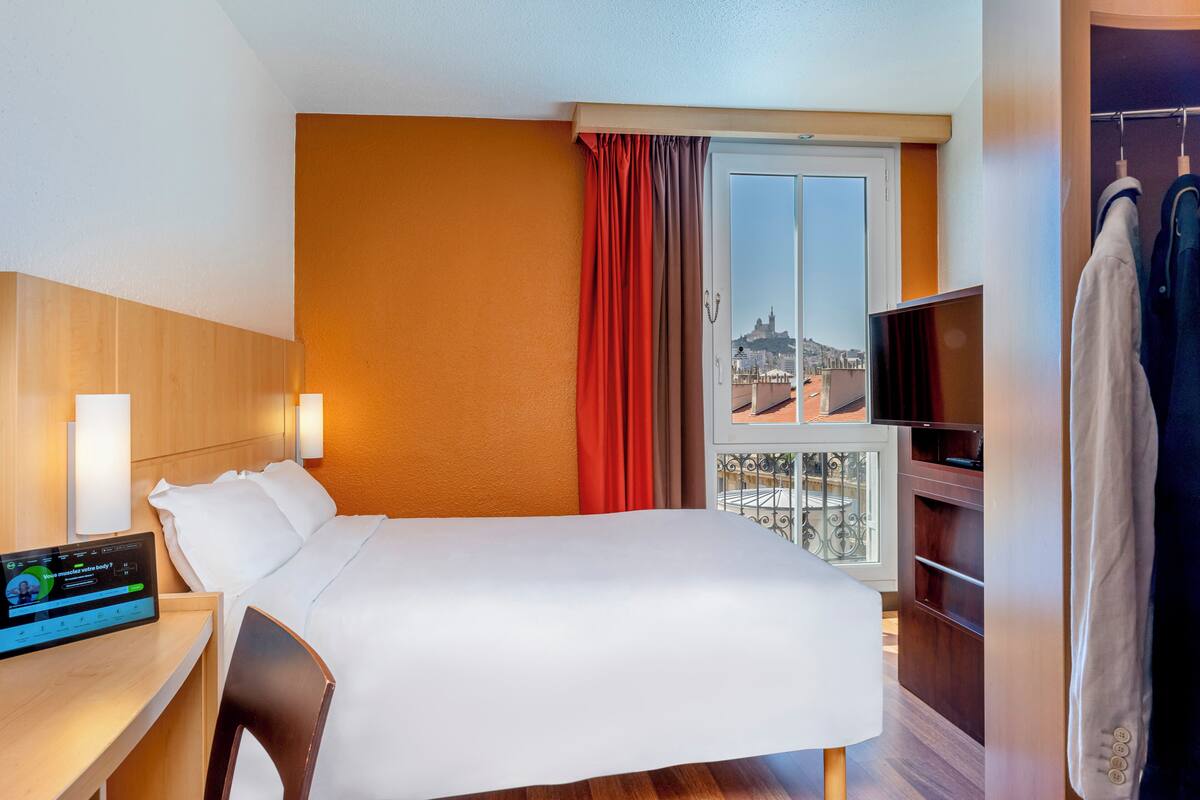 A hotel room with a bed, a desk with a control panel, a TV, a wardrobe, and a window with a view of a cityscape.