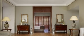 A hotel lobby with a reception desk, two side tables with lamps, a large mirror, and a wooden door leading to a bathroom.