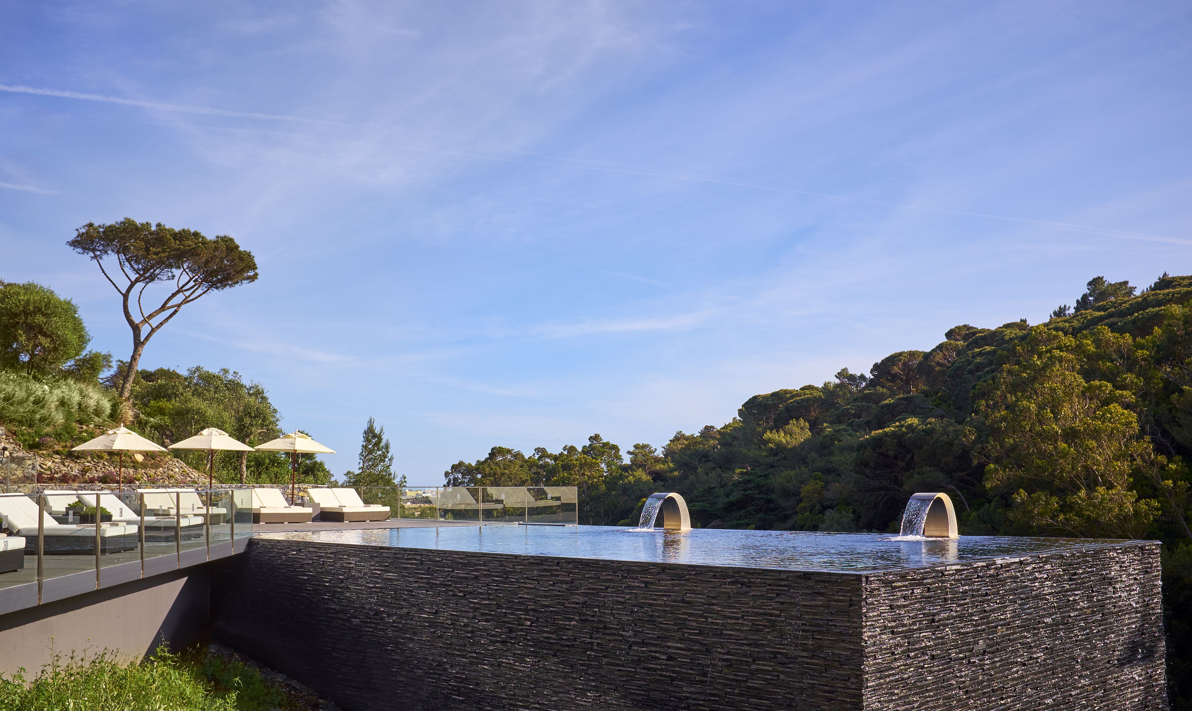 Una piscina con cascada, rodeada de sillas de descanso y sombrillas, con vistas a una ladera.